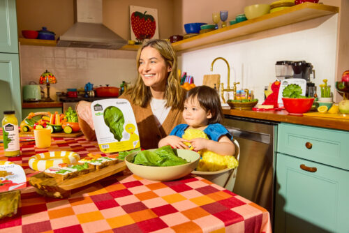 Gotham Greens + Sesame Street collaboration: a mother sits at a table with a package of Big Bird on Romaine lettuce package, while her child sits in a high chair.